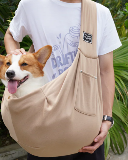 Person holding a corgi in a beige pet sling against a green outdoor background
