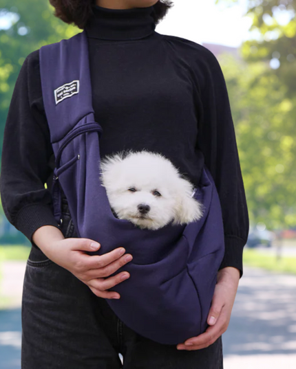 Person holding a small white dog in a purple sling against a blurred green outdoor background