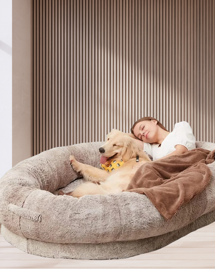 Person lying on a large fluffy pet bed with a dog, in a bright room.