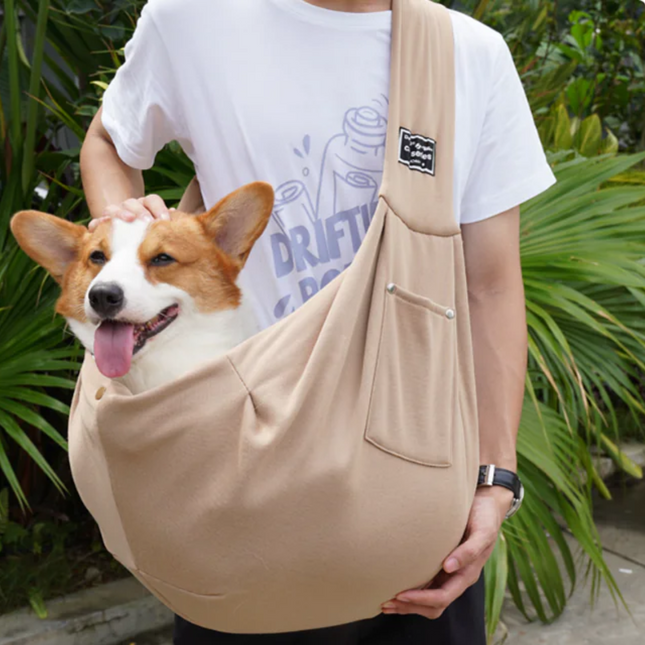 Person holding a corgi in a beige pet sling against a green outdoor background