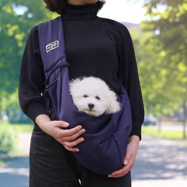 Person holding a small white dog in a purple sling against a blurred green outdoor background