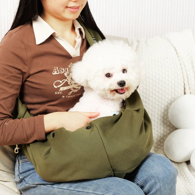 Person holding a small white dog in a green pet sling on a couch.