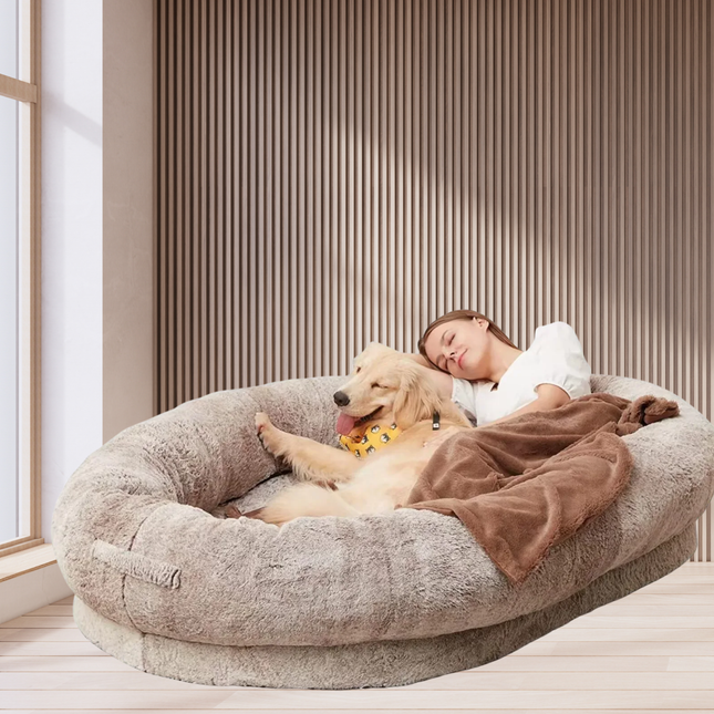 Person lying on a large fluffy pet bed with a dog, in a bright room.